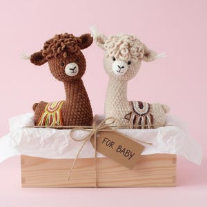 Peut inclure: Deux lamas en crochet, un marron et un crème, sont présentés dans une boîte en bois avec une étiquette « FOR BABY ». Les lamas ont des couvertures de selle décoratives, sur fond rose.
