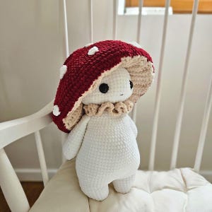 Può includere: Un peluche a forma di fungo fatto a mano all'uncinetto in bianco, bordeaux e beige. Il peluche ha un cappello a forma di fungo con macchie bianche, un colletto a balze e grandi occhi neri. È in piedi su un cuscino bianco.
