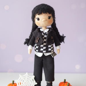 Peut inclure: Poupée crochetée vêtue d'une veste noire, d'un pantalon et d'une chemise blanche à col. La poupée a des tresses noires et se tient devant une toile d'araignée blanche et trois citrouilles oranges. La veste a des boutons marron.