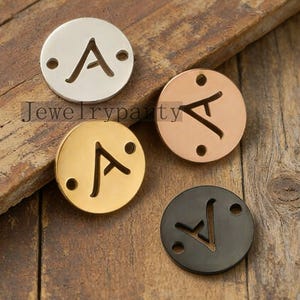 May include: Four metal charms in silver, gold, rose gold, and black. Each charm is a circle with the letter "A" cut out and two small holes. The charms are on a wooden surface. The text "Jewelryparty" is visible.