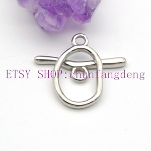 以下が含まれることがあります： 楕円形のループと水平バーを備えたシルバーカラーのトグルクラスプ。クラスプの上部に小さなループがあり、中央に小さなループがあります。「ETSY SHOP：chunfangdeng」というテキストが紫で印刷されています。