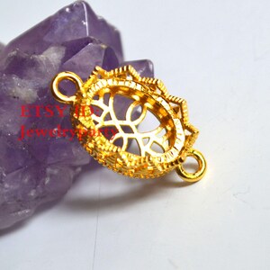 May include: A gold-colored filigree connector with an oval center and decorative edges. The connector has two loops for attaching to other jewelry components. The background is a purple crystal.