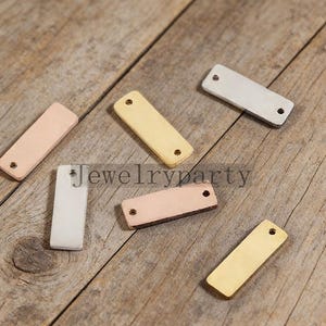 May include: Various rectangular metal jewellery tags in gold, rose gold, and silver. Each tag has two small holes and is displayed on a wooden surface. The text "Jewelryparty" is visible.
