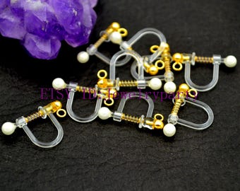 May include: A close-up of a group of clear plastic earring backs with gold spring clips and white pearl accents. The earring backs are designed to be used with clip-on earrings.