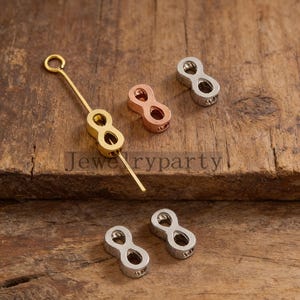 May include: Various metal jewelry findings in gold, rose gold, and silver. The findings are shaped like the number eight, with holes for attachment. The items are displayed on a wooden surface. Measurements are in centimetres.