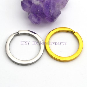 Puede incluir: Dos llaveros, uno plateado y otro dorado, se muestran sobre un fondo blanco con un grupo de cristales morados. El anillo plateado está a la izquierda y el anillo dorado a la derecha. El texto "ETSY ID: Jewelryparty" es visible.