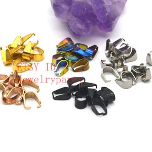 Puede incluir: Un surtido de colgantes para joyería en varios acabados metálicos, incluyendo dorado, iridiscente, plateado, oro rosa y negro. Los colgantes están dispuestos en pequeños montones sobre un fondo blanco, con un cristal púrpura en el fondo. El texto "ETSY ID Jewelrypart" es visible.