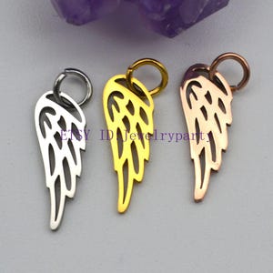 Può includere: Tre ciondoli a forma di ala in metallo argentato, dorato e oro rosa. Ogni ciondolo presenta un design ad ala con un piccolo anello nella parte superiore per il fissaggio ai gioielli. I ciondoli sono esposti su uno sfondo viola chiaro e grigio.