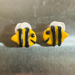 May include: A pair of bee-shaped magnets featuring a bright yellow body with two vertical black stripes, a small black dot for an eye, and a white wing detail on the top. The bees have a glossy finish and are crafted with a rounded, cartoon-like shape.