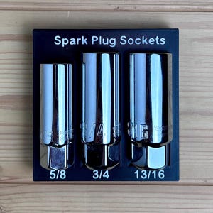 May include: A black rectangular organizer tray labeled Spark Plug Sockets containing three chrome-plated steel spark plug sockets. The sockets are sized 5/8 inch, 3/4 inch, and 13/16 inch, with the sizes clearly printed below each tool.