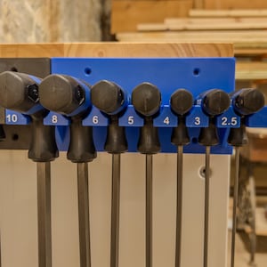 May include: A set of T-handle hex keys with black handles and blue accents, organized in a blue plastic wall-mounted rack. The rack features printed size labels including 3/32, 10, 8, 6, 5, 4, 3, and 2.5, with the metal shafts extending downward.