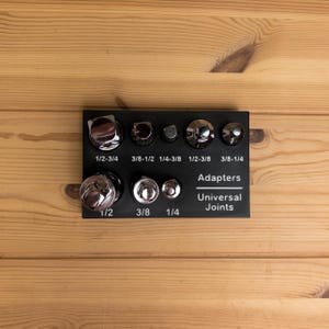 May include: A black rectangular organizer tray holding various chrome-plated socket adapters and universal joints. The tray features white printed labels including 1/2-3/4, 3/8-1/2, 1/4-3/8, 1/2-3/8, 3/8-1/4, 1/2, 3/8, 1/4, Adapters, and Universal Joints.