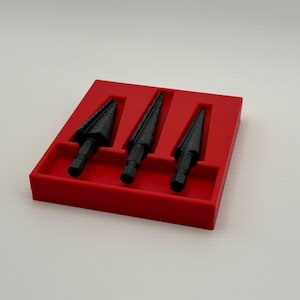 May include: Three black step drill bits are neatly arranged in a bright red plastic tray. The drill bits have a conical shape with multiple stepped cutting edges. The tray provides a secure and organized storage solution for the tools.
