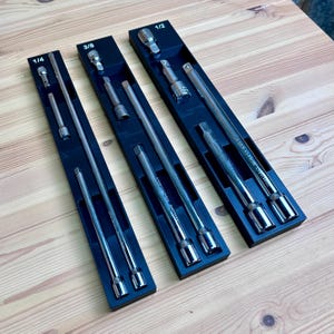 May include: Three black plastic organizers containing silver metal socket wrench extensions. The organizers are labeled 1/4, 3/8, and 1/2 at the top. Each tray holds various lengths of steel extension bars and adapters for mechanical socket sets.