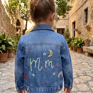 Peut inclure: Une veste en jean bleu avec une broderie au dos du prénom Mimi en vert clair, entourée de motifs cousus incluant un croissant de lune jaune, de petites étoiles, des fleurs violettes, une paire de cerises rouges et un petit arc-en-ciel pastel.