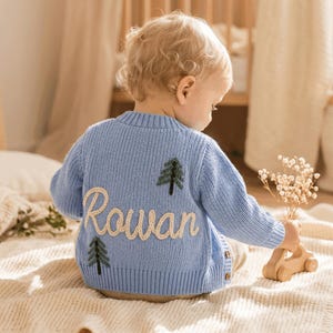 Pode incluir: Um cardigan de malha azul claro com o nome "Rowan" bordado em fio creme. Duas aplicações de árvores verdes estão nas costas. O cardigan tem fecho de botão e é projetado para uma criança.