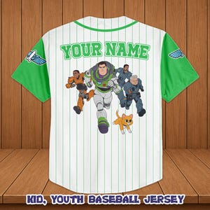 Puede incluir: Camiseta de b&eacute;isbol blanca con mangas y cuello verdes, con un gr&aacute;fico de dibujos animados de Buzz Lightyear y otros personajes. La camiseta tiene rayas verticales y el texto "YOUR NAME" en verde. El texto inferior dice "KID, YOUTH BASEBALL JERSEY".