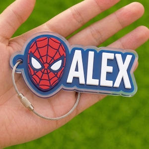 May include: A custom keychain featuring a red and blue Spider-Man mask graphic next to the name ALEX in bold white letters on a dark blue background. The design is encased in clear acrylic and attached to a flexible silver-toned metal wire loop.