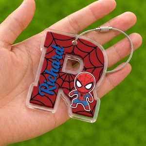 May include: A clear acrylic keychain shaped like the letter R, featuring a red background with a black spiderweb pattern. The name Richard is printed in blue script, and a small cartoon Spider-Man figure is attached, with a silver wire loop.