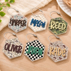 May include: Six hexagonal acrylic keychains with metal wire loops. Each features a unique pattern including camouflage, cow print, and checkerboard. The names Harper, Mason, Reed, Oliver, Jaxon, and Kade are displayed in raised, colorful 3D lettering.