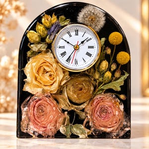 May include: An arched black resin desk clock featuring embedded dried flowers including pink and yellow roses, yellow billy balls, and a dandelion seed head. The clock face is white with black Roman numerals and gold-tone trim.