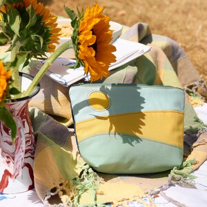 May include: A zipped pouch featuring a landscape design with curved panels of light blue and mustard yellow fabric, including a circular yellow sun appliqué. The pouch has a dark blue zip closure and visible stitching along the fabric seams.