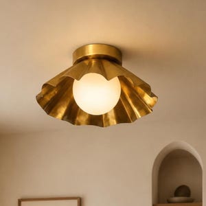 May include: A flush mount ceiling light fixture featuring a metallic gold, scalloped, fan-shaped shade. A central, spherical, frosted white glass globe sits within the center of the brass-toned metal shade, emitting a soft, warm, diffused light.
