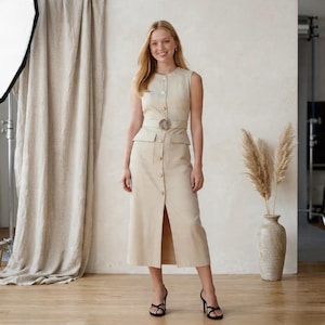 Può includere: Abito beige senza maniche con cintura abbinata e bottoni dorati. L'abito ha una gonna al ginocchio con uno spacco frontale. La modella indossa sandali neri con tacco alto. Lo sfondo comprende una tenda beige e un vaso con piante decorative.