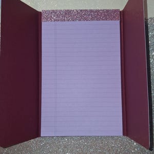 Puede incluir: Un bloc de notas con papel rayado de color morado claro y un encabezado de purpurina oro rosa, dentro de una cubierta plegable de color granate oscuro. El interior de la cubierta es granate s&oacute;lido y el exterior tiene un acabado de purpurina.