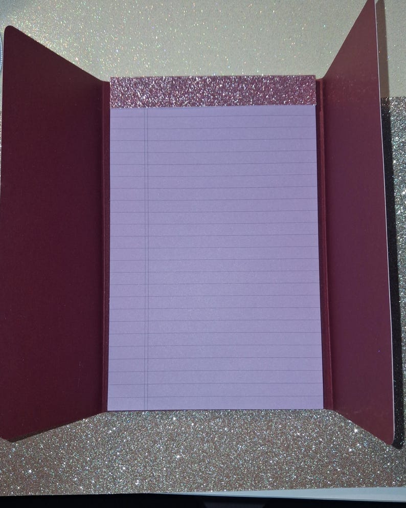 Puede incluir: Un bloc de notas con papel rayado de color morado claro y un encabezado de purpurina oro rosa, dentro de una cubierta plegable de color granate oscuro. El interior de la cubierta es granate s&oacute;lido y el exterior tiene un acabado de purpurina.