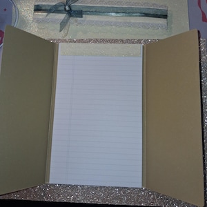 Puede incluir: Una carpeta de papeler&iacute;a de tres pliegues con un exterior de purpurina color champ&aacute;n. El centro cuenta con un bloc de notas blanco con l&iacute;neas y un borde superior brillante. La solapa superior est&aacute; decorada con una cinta verde azulado, un lazo transparente y un borde de encaje color crema.