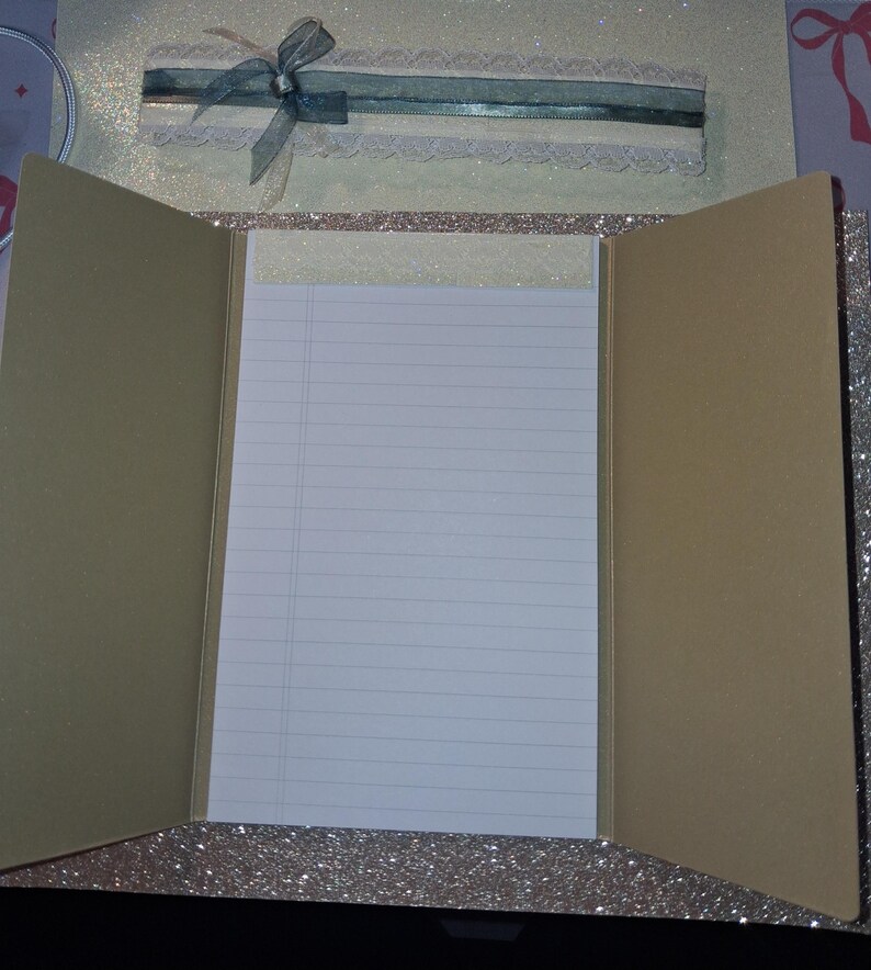 Puede incluir: Una carpeta de papeler&iacute;a de tres pliegues con un exterior de purpurina color champ&aacute;n. El centro cuenta con un bloc de notas blanco con l&iacute;neas y un borde superior brillante. La solapa superior est&aacute; decorada con una cinta verde azulado, un lazo transparente y un borde de encaje color crema.