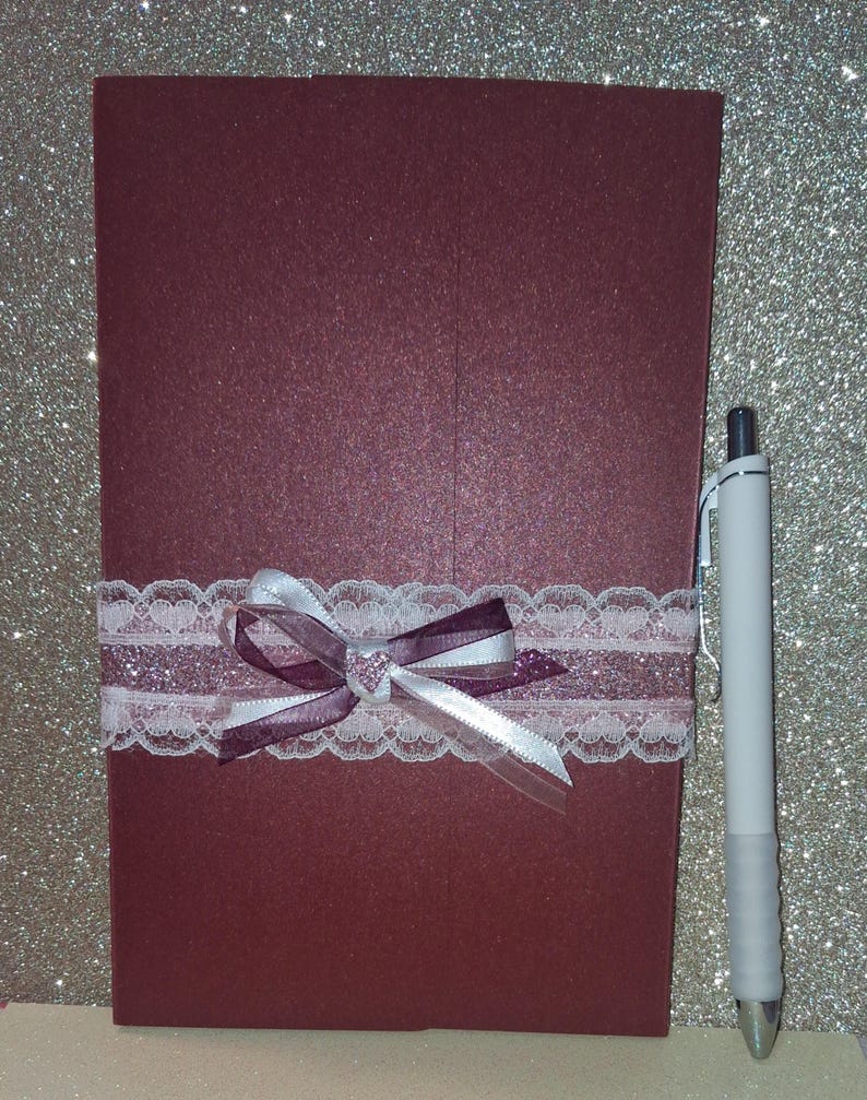 Puede incluir: Una carpeta rectangular de cartulina color burdeos oscuro con una banda horizontal de encaje blanco sobre una tira rosa brillante. Un lazo decorativo de cinta blanca y burdeos se sit&uacute;a en el centro, acompa&ntilde;ado de un bol&iacute;grafo blanco retr&aacute;ctil.