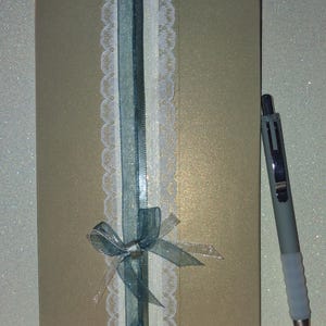 Puede incluir: Una tarjeta rectangular de tono dorado con una franja decorativa vertical que presenta un borde de encaje blanco, una cinta azul transparente y una cinta plateada fina atada en un lazo en la parte inferior. Un bol&iacute;grafo gris est&aacute; a la derecha.