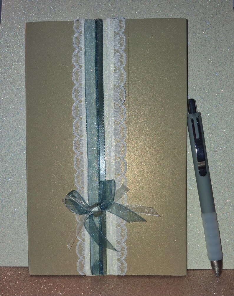Puede incluir: Una tarjeta rectangular de tono dorado con una franja decorativa vertical que presenta un borde de encaje blanco, una cinta azul transparente y una cinta plateada fina atada en un lazo en la parte inferior. Un bol&iacute;grafo gris est&aacute; a la derecha.