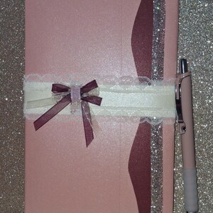 Puede incluir: Un cuaderno rectangular de color rosa claro con una franja vertical de color granate oscuro y purpurina plateada en el lado derecho. Una cinta de encaje blanco envuelve el centro horizontalmente, con un peque&ntilde;o lazo granate y un detalle transparente.