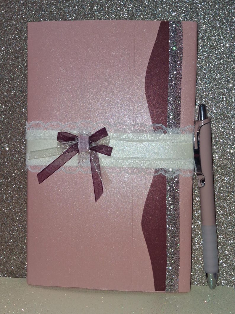 Puede incluir: Un cuaderno rectangular de color rosa claro con una franja vertical de color granate oscuro y purpurina plateada en el lado derecho. Una cinta de encaje blanco envuelve el centro horizontalmente, con un peque&ntilde;o lazo granate y un detalle transparente.