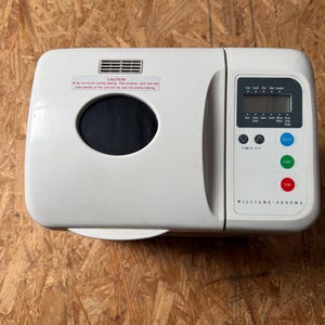 May include: A white Williams-Sonoma bread maker with a digital display and control buttons. The machine has a viewing window and a warning label that reads "CAUTION". The bread maker is on a wooden surface.