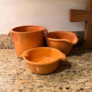 May include: A set of three glossy orange ceramic mixing bowls of varying sizes, each featuring two small side handles and a smooth, rounded design. The bowls are arranged in a cluster, showcasing their vibrant, solid orange finish and functional, open-top structure for kitchen use.