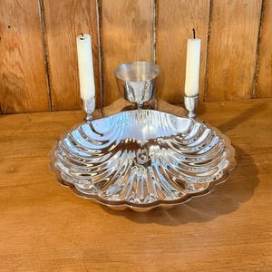 May include: A silver-toned metal shell-shaped dish featuring a central small bowl and two integrated candle holders, each containing a white taper candle. The dish has a scalloped edge and a reflective, polished surface with radial ridges.