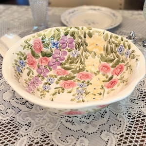 May include: A round ceramic bowl with a scalloped rim, featuring a hand-painted floral pattern of pink roses, purple clusters, and yellow blossoms interspersed with green leaves against a white background, creating a decorative serving piece.