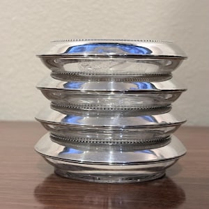 May include: A stack of four circular coasters, each featuring a clear glass center surrounded by a polished silver-tone metal rim. The metal edges are decorated with a fine beaded texture along the inner border, creating a uniform appearance.