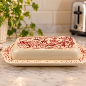 May include: A cream-colored ceramic butter dish with a rectangular lid and base. The lid features a detailed red transferware print depicting a horse-drawn carriage with passengers, while the base has a decorative red border pattern.