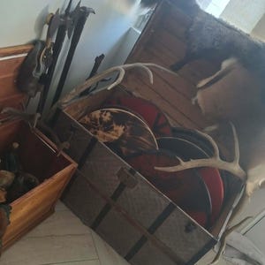 May include: A large wooden chest with metal corner reinforcements containing several round shields with red and black painted designs, accented by deer antlers. Beside it sits a smaller wooden box holding dark axes and a leather pouch.