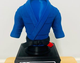 May include: A blue martial arts gi top with a black belt featuring a red tip, mounted on a black rectangular base. The base displays a white plaque with the Japanese kanji for Jiu-Jitsu inside a red circle next to the name Eduardo in white text.