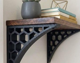 May include: A dark wood shelf supported by two black metal brackets featuring a geometric honeycomb pattern. The shelf holds a small gray pot with a succulent, a stack of three books with light-colored covers, and a gold-toned wire sculpture.