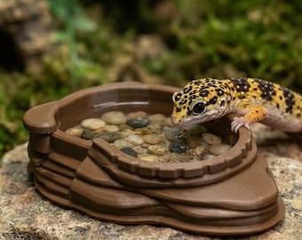 May include: A brown, layered, circular water dish filled with small, smooth, multicolored pebbles. A yellow and black spotted leopard gecko rests its front feet on the rim of the dish, looking into the water.