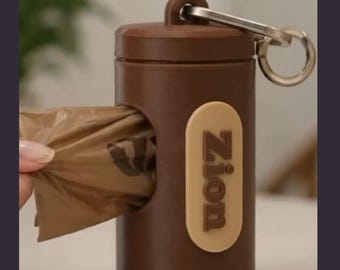 May include: A cylindrical brown plastic waste bag dispenser featuring a metal carabiner clip at the top. A beige oval label on the front displays the name Zion in brown lettering. A brown plastic waste bag is partially pulled through the side opening.