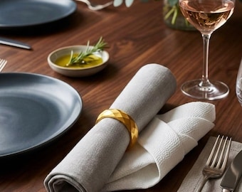May include: A gold-colored napkin ring with a faceted geometric surface holding a rolled light gray fabric napkin. The napkin ring features a polished metallic finish and a multi-sided design, resting on a dark wooden table with place settings.