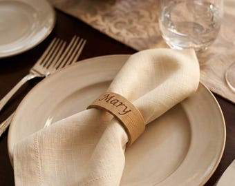 May include: A light beige fabric napkin folded and secured with a smooth, circular wooden napkin ring. The wooden ring features the name Mary engraved in a cursive script font. The napkin is placed on a round, off-white ceramic dinner plate.